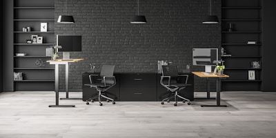 A modern office space with two workstations featuring adjustable desks and ergonomic chairs. The background includes a dark brick wall with symmetrical black shelves, adorned with minimalistic decor such as books, framed art, and plants. The lighting is provided by sleek pendant lamps.