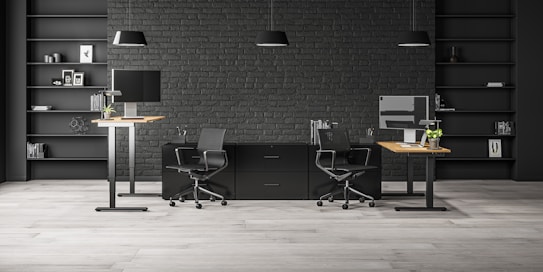 A modern office space with two workstations featuring adjustable desks and ergonomic chairs. The background includes a dark brick wall with symmetrical black shelves, adorned with minimalistic decor such as books, framed art, and plants. The lighting is provided by sleek pendant lamps.