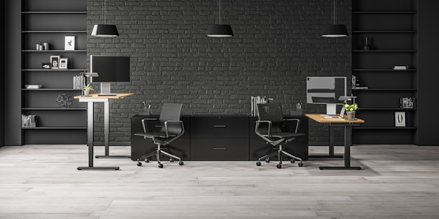A modern office space with two workstations featuring adjustable desks and ergonomic chairs. The background includes a dark brick wall with symmetrical black shelves, adorned with minimalistic decor such as books, framed art, and plants. The lighting is provided by sleek pendant lamps.