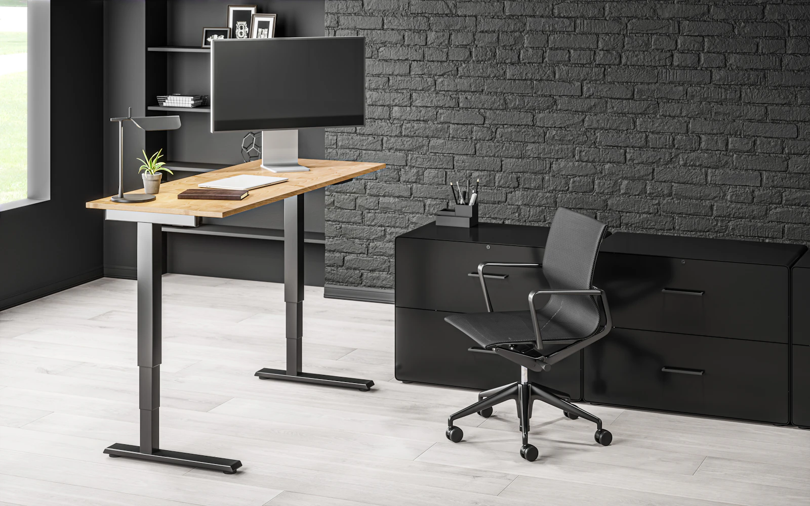 A height-adjustable standing desk with a monitor on a riser, desk lamp, and an ergonomic mesh office chair in a modern home office