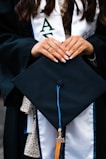 A person wearing a graduation gown holds a graduation cap with a blue and gold tassel marked '2021'. The person's hands are adorned with neatly manicured nails and a small ring.