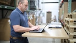A man in a blue polo shirt standing at a wooden standing desk with a laptop in a warehouse setting. He appears focused and is typing on the keyboard. To the right, there are stacked pallets and a scale on the desk.