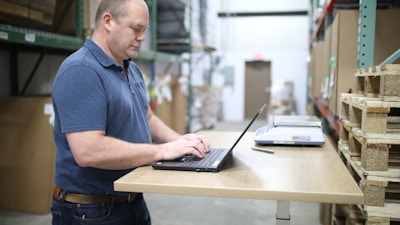 A man in a blue polo shirt standing at a wooden standing desk with a laptop in a warehouse setting. He appears focused and is typing on the keyboard. To the right, there are stacked pallets and a scale on the desk.