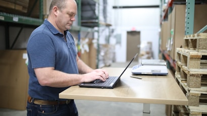 A man in a blue polo shirt standing at a wooden standing desk with a laptop in a warehouse setting. He appears focused and is typing on the keyboard. To the right, there are stacked pallets and a scale on the desk.