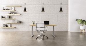 A minimalist office space with a sleek wooden desk and a modern chair set against a concrete wall. Three black pendant lights hang symmetrically above the desk. To the left, open wooden shelves are adorned with books, decorative items, and framed pictures. A potted plant adds a touch of greenery to the right side of the room.