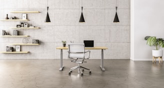 A minimalist office space with a sleek wooden desk and a modern chair set against a concrete wall. Three black pendant lights hang symmetrically above the desk. To the left, open wooden shelves are adorned with books, decorative items, and framed pictures. A potted plant adds a touch of greenery to the right side of the room.