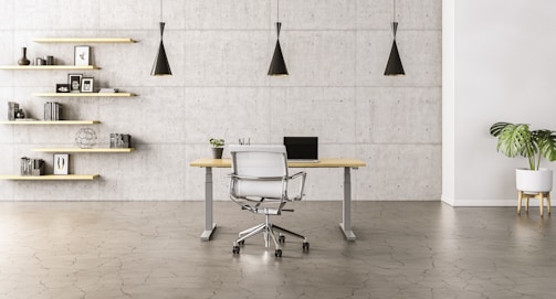 A minimalist office space with a sleek wooden desk and a modern chair set against a concrete wall. Three black pendant lights hang symmetrically above the desk. To the left, open wooden shelves are adorned with books, decorative items, and framed pictures. A potted plant adds a touch of greenery to the right side of the room.