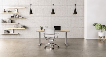 A minimalist office space with a sleek wooden desk and a modern chair set against a concrete wall. Three black pendant lights hang symmetrically above the desk. To the left, open wooden shelves are adorned with books, decorative items, and framed pictures. A potted plant adds a touch of greenery to the right side of the room.
