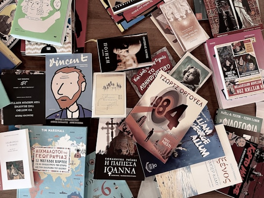 A collection of various books spread out on a wooden surface. The books have diverse cover designs, including illustrations and text in different languages. Prominent titles include 'Vincent,' '1984,' and 'Ioanna.' The covers feature a mix of artistic styles ranging from graphic and comic styles to classical depictions.