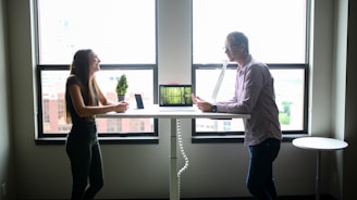 Two people engaging in conversation while standing across from each other at a desk in a well-lit office environment. A computer and a small potted plant are placed on the desk. The scene is set against large windows with an urban cityscape visible outside.