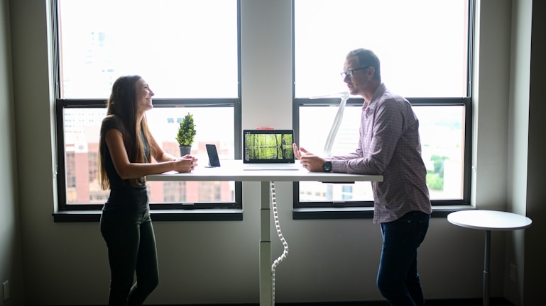 Two people engaging in conversation while standing across from each other at a desk in a well-lit office environment. A computer and a small potted plant are placed on the desk. The scene is set against large windows with an urban cityscape visible outside.