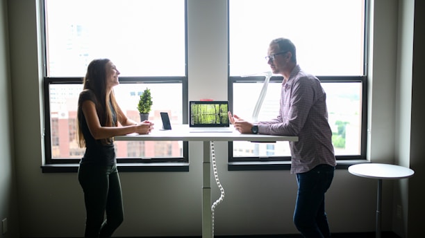 Two people engaging in conversation while standing across from each other at a desk in a well-lit office environment. A computer and a small potted plant are placed on the desk. The scene is set against large windows with an urban cityscape visible outside.