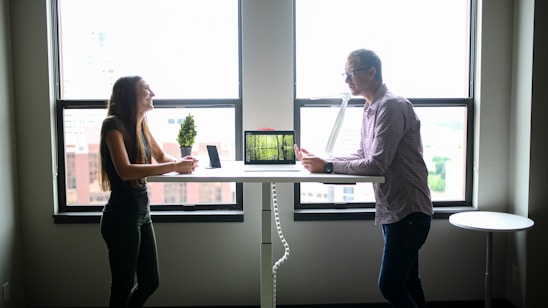 Two people engaging in conversation while standing across from each other at a desk in a well-lit office environment. A computer and a small potted plant are placed on the desk. The scene is set against large windows with an urban cityscape visible outside.