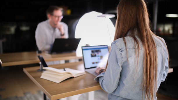 A young woman with long hair, wearing a blue denim jacket, works on a laptop at a table in a dimly lit room. A bright light source, possibly a lamp, is positioned in front of her. On the table, there is an open book and a microphone. In the background, another person is visible, also using a laptop.