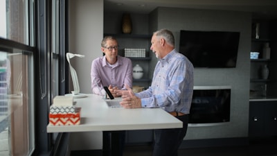 Two men are engaged in a conversation at a standing desk in a modern office environment. One man, wearing glasses and a checkered shirt, is seated and appears to be listening attentively, while the other man, dressed in a similar shirt, is leaning on the desk, gesturing with his hands as he speaks. The room features a large flat-screen TV on the wall, a bookshelf with decorative items, and large windows on one side.