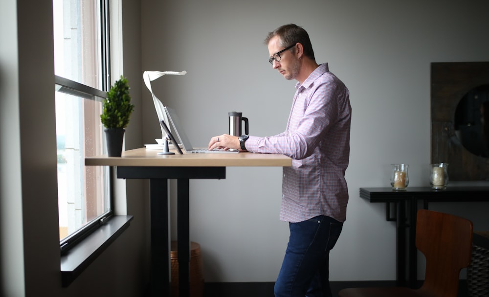 man in pink dress shirt and blue denim jeans standing beside brown wooden table