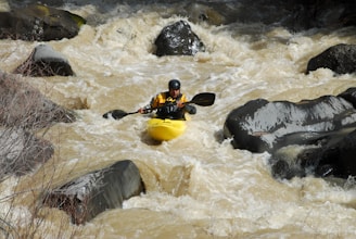A person is kayaking through rough whitewater rapids surrounded by large rocks. The water appears turbulent and fast-moving, suggesting a challenging and adventurous environment. The kayaker is wearing a helmet and a yellow kayak jacket.