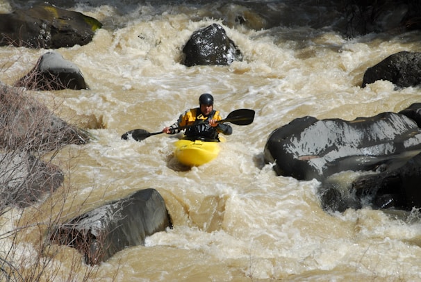 A person is kayaking through rough whitewater rapids surrounded by large rocks. The water appears turbulent and fast-moving, suggesting a challenging and adventurous environment. The kayaker is wearing a helmet and a yellow kayak jacket.