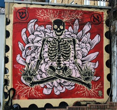 A street art mural featuring a skeleton in a crossed-leg, meditative pose against a vibrant red background. The skeleton's head is adorned with thin, branch-like antlers, and it sits atop a design resembling flower petals, executed in light pink and white hues. The skeleton's legs and hands hold torch-like elements emitting roots, blending into the flower motif. Black circular patterns frame the edges of the mural, which displays weathered portions and urban graffiti tags.