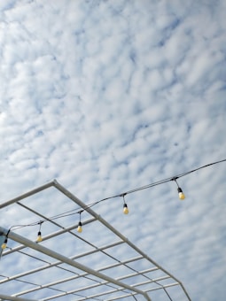 A cloudy sky serves as the backdrop with scattered small, fluffy clouds. In the foreground, a metallic grid structure is visible with string lights hanging across it. The bulbs are evenly spaced, creating a line parallel to the bars of the structure.