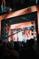 A vibrant concert scene featuring a large stage with bright, colorful LED lights. Several performers are on stage, while a crowd of people watches. A large camera crane is visible, capturing the event.