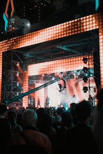 A vibrant concert scene featuring a large stage with bright, colorful LED lights. Several performers are on stage, while a crowd of people watches. A large camera crane is visible, capturing the event.