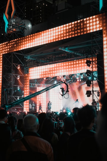 A vibrant concert scene featuring a large stage with bright, colorful LED lights. Several performers are on stage, while a crowd of people watches. A large camera crane is visible, capturing the event.