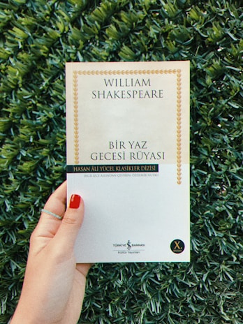 A person is holding a book with a white cover, titled 'William Shakespeare - Bir Yaz Gecesi Rüyası,' surrounded by green foliage. The person's fingernails are painted red, and they are wearing a thin ring.
