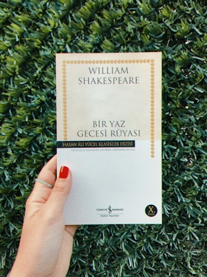 A person is holding a book with a white cover, titled 'William Shakespeare - Bir Yaz Gecesi Rüyası,' surrounded by green foliage. The person's fingernails are painted red, and they are wearing a thin ring.