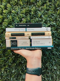 A hand with a wristband holds a stack of books against a lush green background. The books include titles by authors like George Orwell, Paulo Coelho, and Jane Austen. The grass is dense and vibrant, contrasting with the varied shades of the book covers.