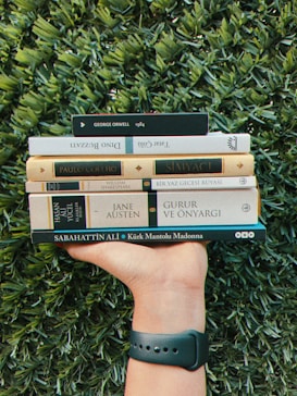 A hand with a wristband holds a stack of books against a lush green background. The books include titles by authors like George Orwell, Paulo Coelho, and Jane Austen. The grass is dense and vibrant, contrasting with the varied shades of the book covers.