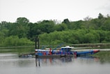 A blue and red dredging machine floats on a calm, reflective body of water, surrounded by lush green trees and vegetation. The machine is equipped with multiple attachments and appears to be in operation, with various mechanical parts visible.