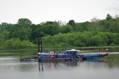 A blue and red dredging machine floats on a calm, reflective body of water, surrounded by lush green trees and vegetation. The machine is equipped with multiple attachments and appears to be in operation, with various mechanical parts visible.