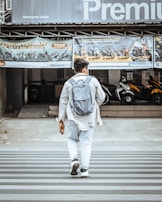 A person wearing a light jacket and carrying a backpack walks across a zebra crossing. The background features a motorcycle dealership displaying bikes and promotional banners.