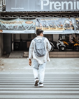 A person wearing a light jacket and carrying a backpack walks across a zebra crossing. The background features a motorcycle dealership displaying bikes and promotional banners.