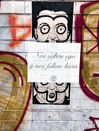 A painted mural on a brick wall featuring a stylized black and white face with exaggerated eyes. The mural is partially covered by a white paper sign with elegant cursive writing in Spanish, reading 'Nos sobra ego y nos faltan luces.' The wall is also adorned with graffiti consisting of red and gold shapes and squiggles.