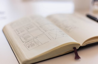An open notebook with sketches and wireframes drawn on its pages, resting on a surface with a soft focus in the background. The pages display various design layouts and diagrams.