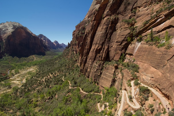A rugged landscape featuring a steep, towering rock face on the right side, with a narrow, winding hiking trail traversing the slope. The foreground showcases lush green vegetation, while distant rocky mountains rise against a clear blue sky.