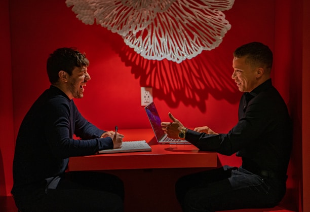 Two people are sitting at a red table, engaged in a lively discussion. One person is writing in a notebook and appears to be laughing, while the other person is gesturing with his hand and working on a laptop. A decorative white hanging piece casts intricate shadows on the red wall behind them.