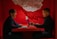 Two people are sitting at a red table, engaged in a lively discussion. One person is writing in a notebook and appears to be laughing, while the other person is gesturing with his hand and working on a laptop. A decorative white hanging piece casts intricate shadows on the red wall behind them.