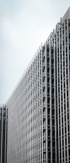 A tall, modern building with a grid-like exterior made of glass and metal. The building's design features numerous evenly spaced vertical and horizontal lines. The top part of the building displays the word 'BERLIN'. The sky in the background is overcast.