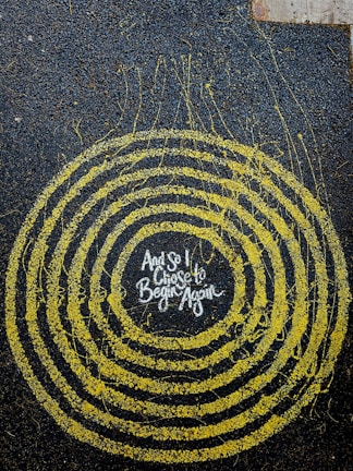 A series of concentric yellow circles on a dark textured background with the words 'And so I chose to begin again' in the center. The circles appear to be made with a splatter effect, creating an abstract and artistic look.