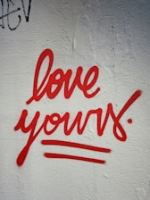 Red graffiti text reading 'love yours' is painted on a textured white wall. The handwriting style is bold and cursive, with underlines emphasizing the message.