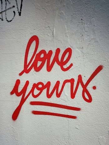 Red graffiti text reading 'love yours' is painted on a textured white wall. The handwriting style is bold and cursive, with underlines emphasizing the message.