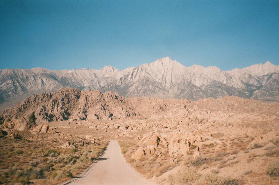 The image depicts a desert landscape with a dirt road winding through it, leading towards a majestic mountain range in the distance. The mountains are covered in light brown rocks and appear to be covered in snow at their base. The sky above is a clear blue, providing a striking contrast to the earthy tones of the mountains and the road.

The perspective of the image is from ground level, looking down towards the mountains, giving a sense of depth and scale. The colors in the photo are predominantly browns and blues, with some greens from vegetation or grass patches on either side of the road.