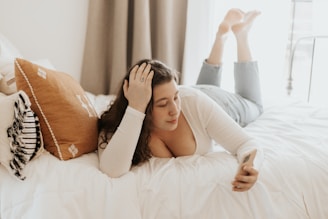A person is lying on a bed with their legs bent at the knees, holding a smartphone. They appear relaxed, dressed in a white top and jeans, and are surrounded by pillows including a prominent orange one.
