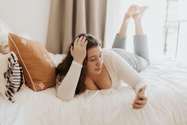 A person is lying on a bed with their legs bent at the knees, holding a smartphone. They appear relaxed, dressed in a white top and jeans, and are surrounded by pillows including a prominent orange one.