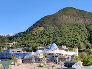A theme park entrance is surrounded by lush green hills and a clear blue sky. Cable cars are visible ascending the hillside. The area is bustling with colorful decorations and signs, reflecting a lively environment.