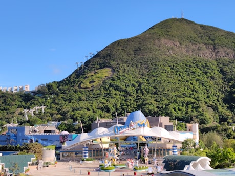 A theme park entrance is surrounded by lush green hills and a clear blue sky. Cable cars are visible ascending the hillside. The area is bustling with colorful decorations and signs, reflecting a lively environment.