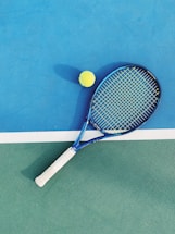 A tennis racket with a blue frame and white grip lies on a two-toned tennis court. Next to it is a yellow tennis ball. The court features a vibrant blue section and a green section divided by a white line.